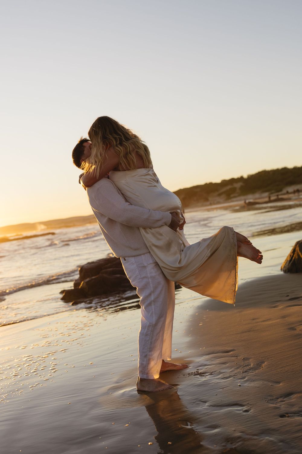 A couple embraces on a beach at sunset with waves lapping at their feet, creating a romantic silhouette against golden light.