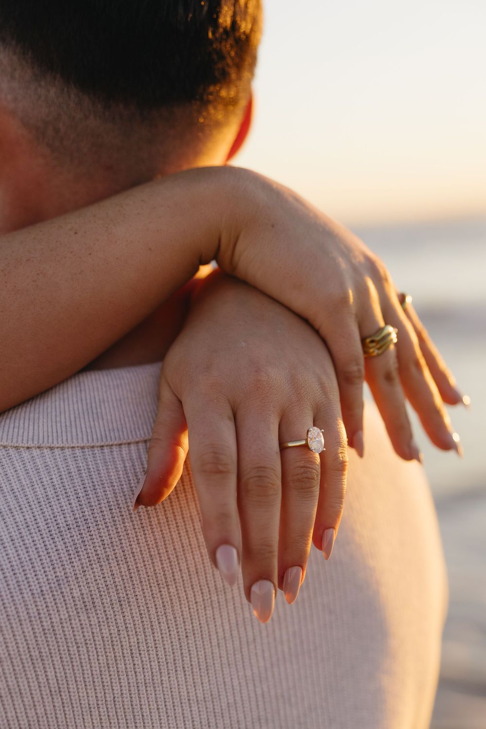 A close-up of hands embracing during sunset, showcasing an engagement ring and manicured nails in warm golden light.