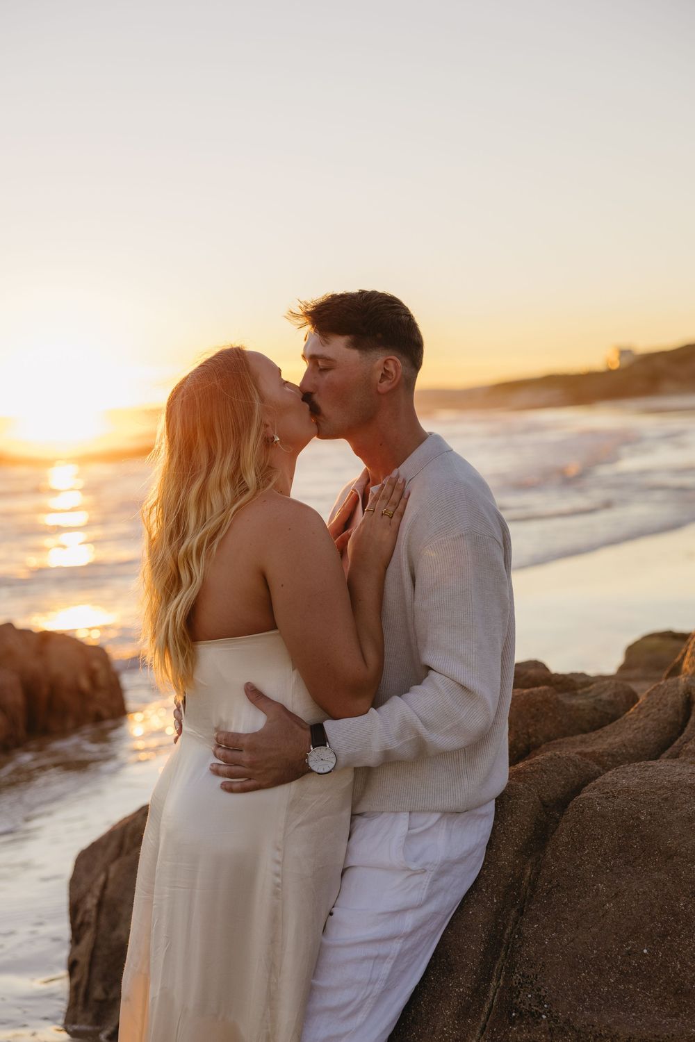 A couple shares romantic kisses on a rocky beach at sunset while waves crash behind them in golden light.