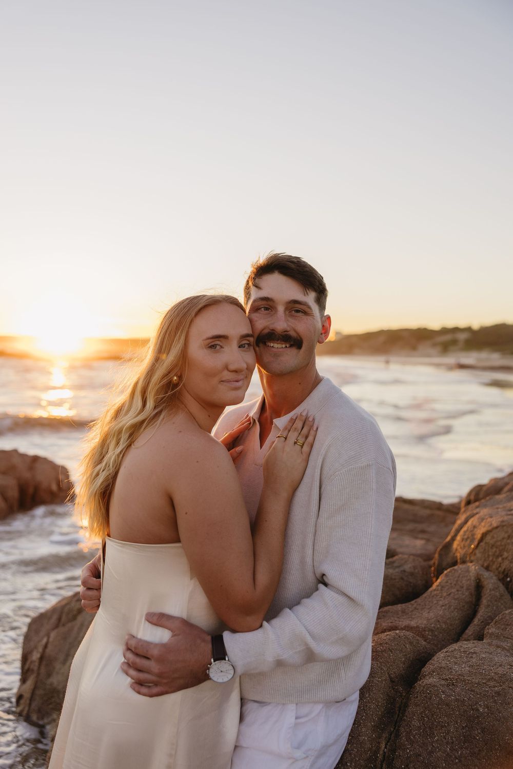 Romantic sunset beach engagement photos showing couple embracing on rocky coastline at golden hour.