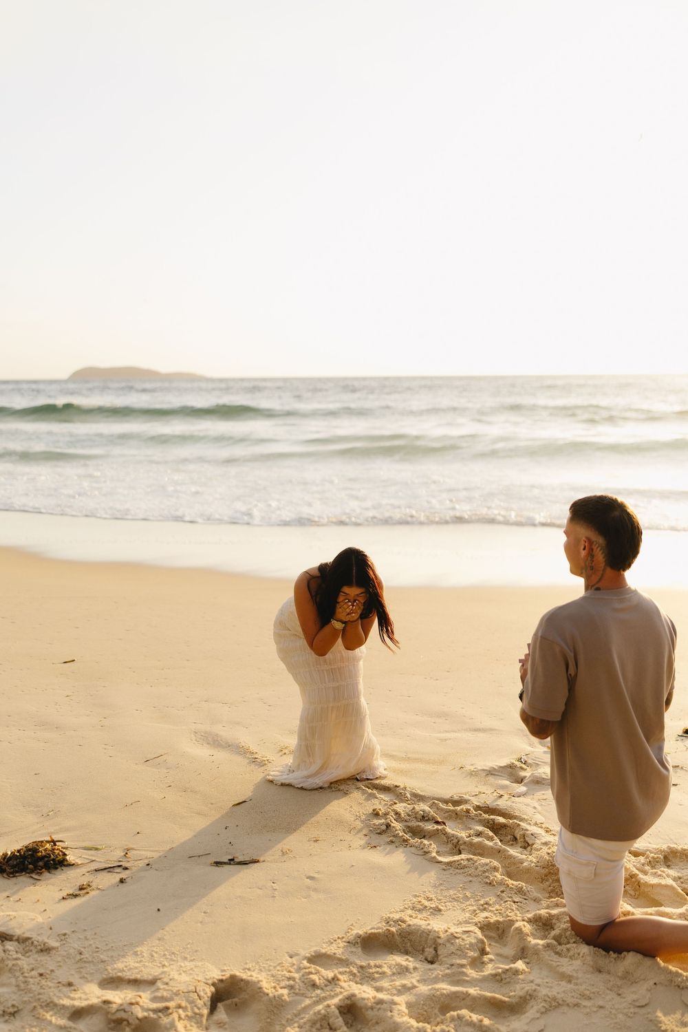 A couple shares an emotional moment during sunset on a sandy beach with gentle waves in the background.