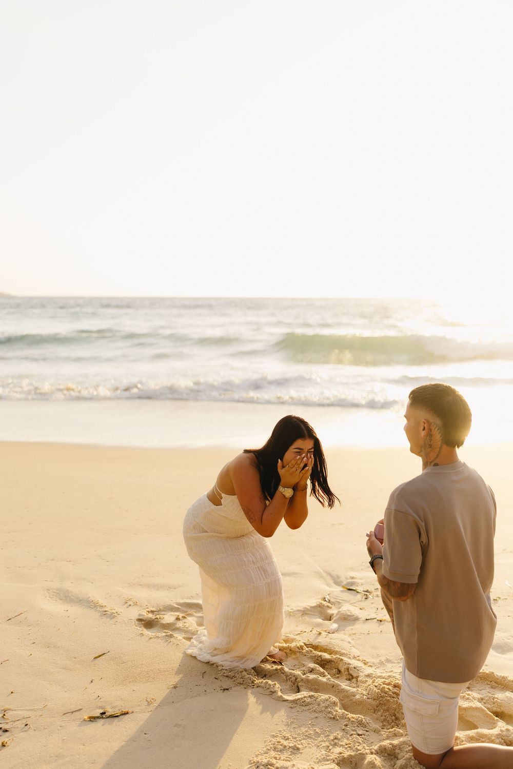 A couple shares a romantic moment during sunset on a sandy beach with ocean waves in the background.