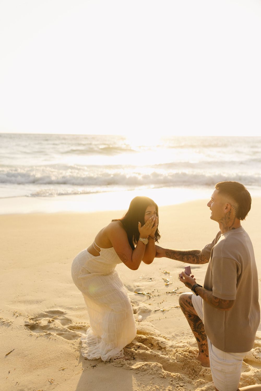 A romantic beach proposal at sunset with two people on the sand as waves roll in the background.