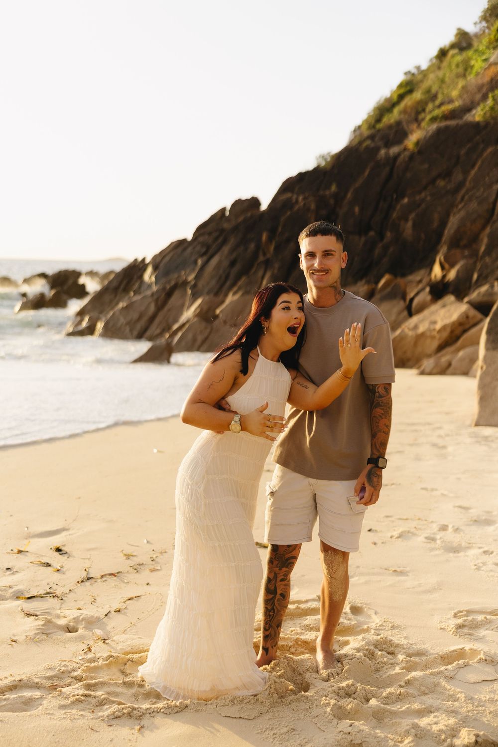 A couple shares a romantic moment on a sandy beach with rocky cliffs in the background during golden hour.