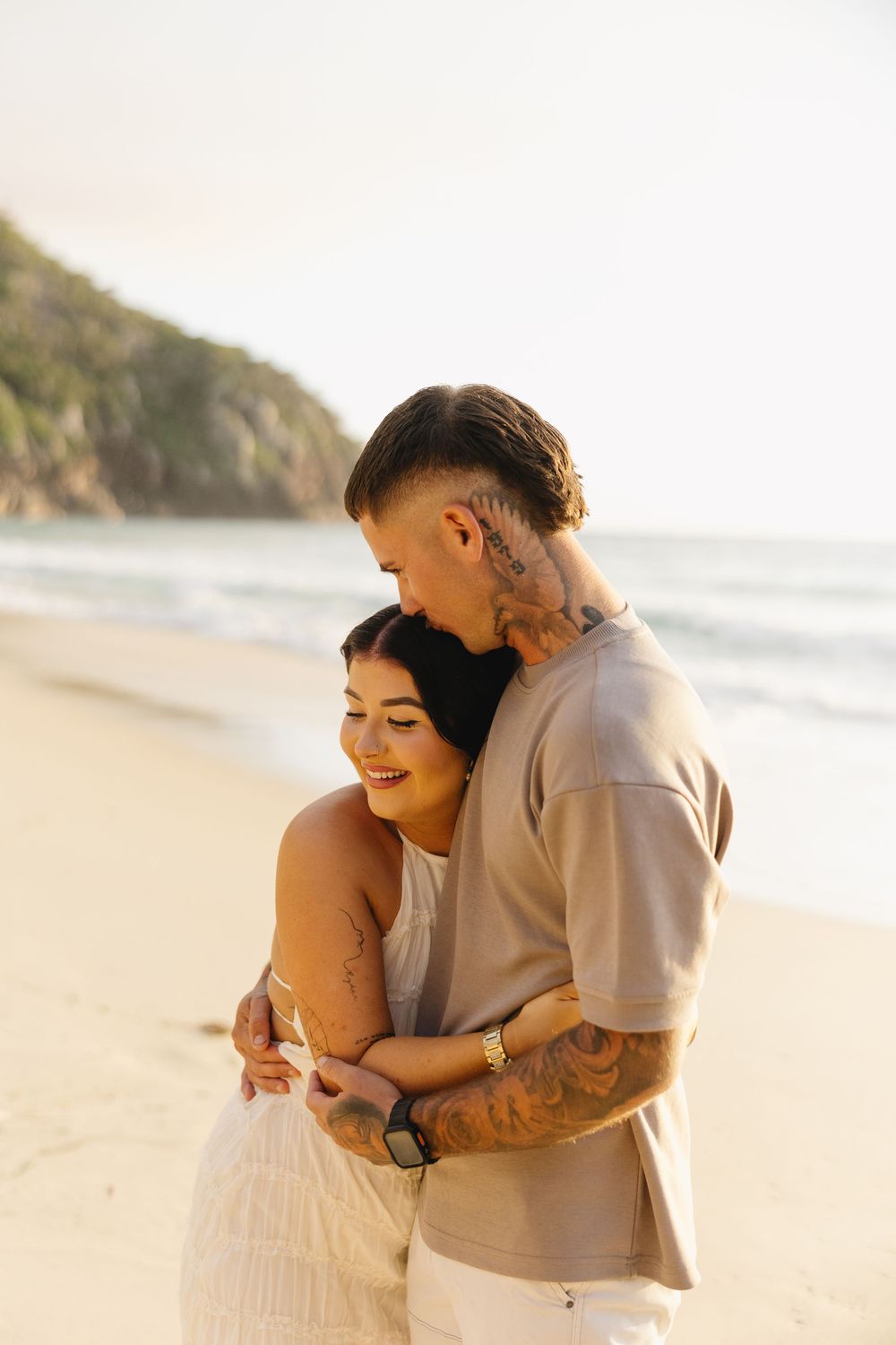A couple shares an intimate moment together on a sunny beach with waves and cliffs in the background.