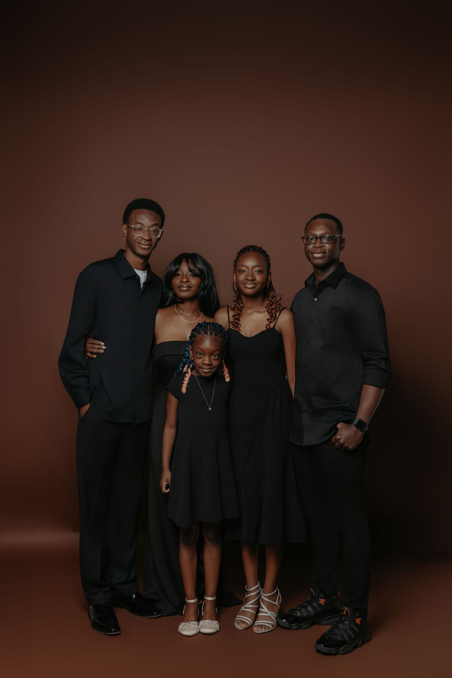 A group of people wearing black clothing pose together in a series of stylish studio portraits against a dark brown background.