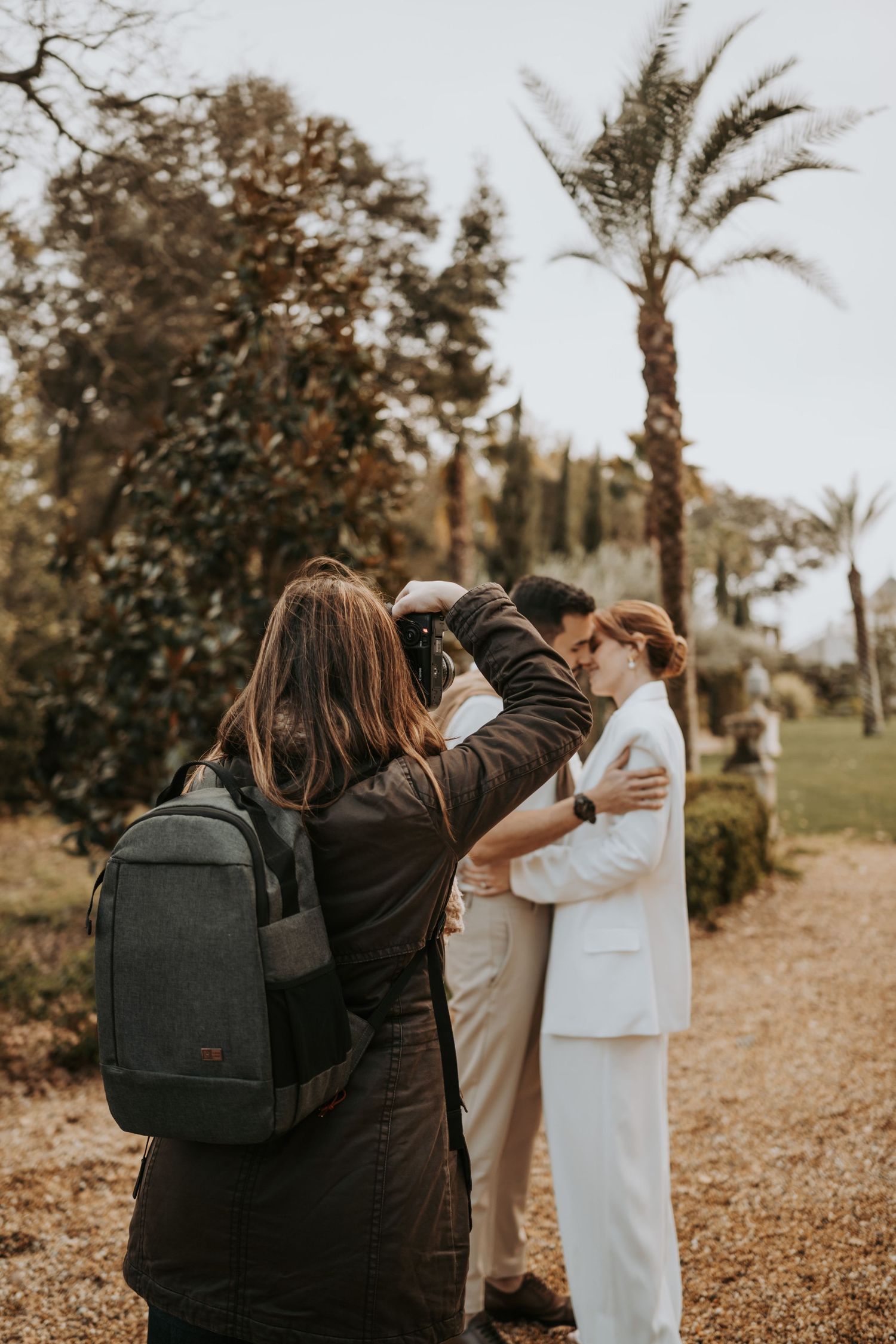 Photographer with backpack captures couple in casual attire under palm trees on a rustic outdoor path.