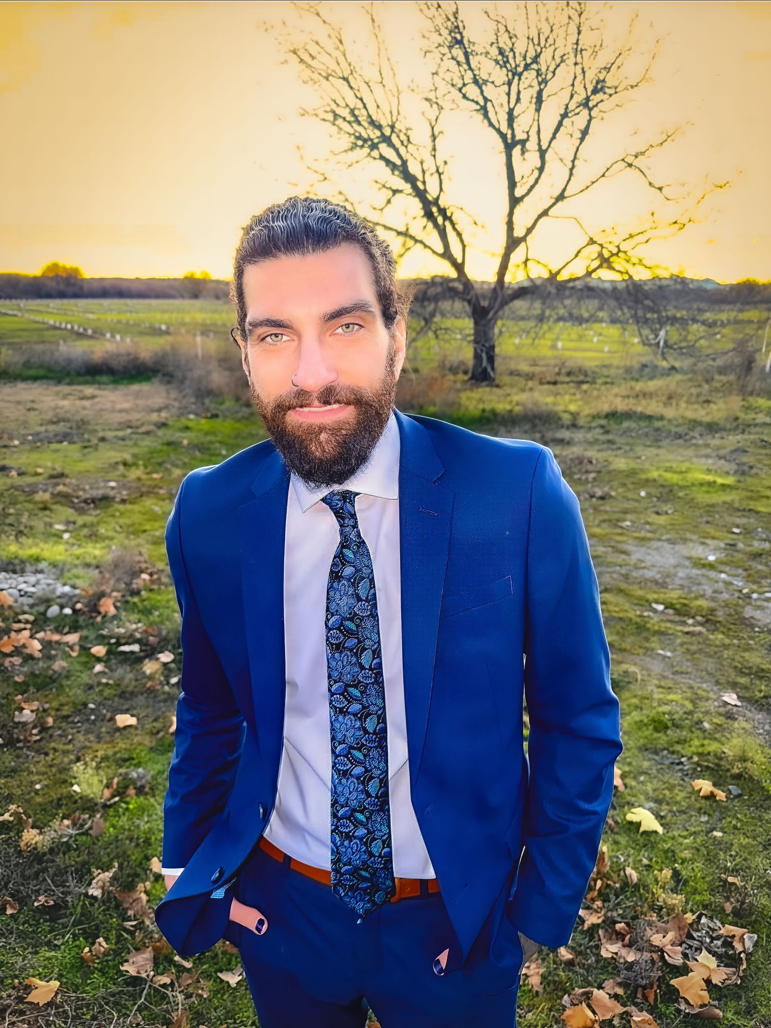 Person in tailored navy suit standing in field with bare tree at golden hour.