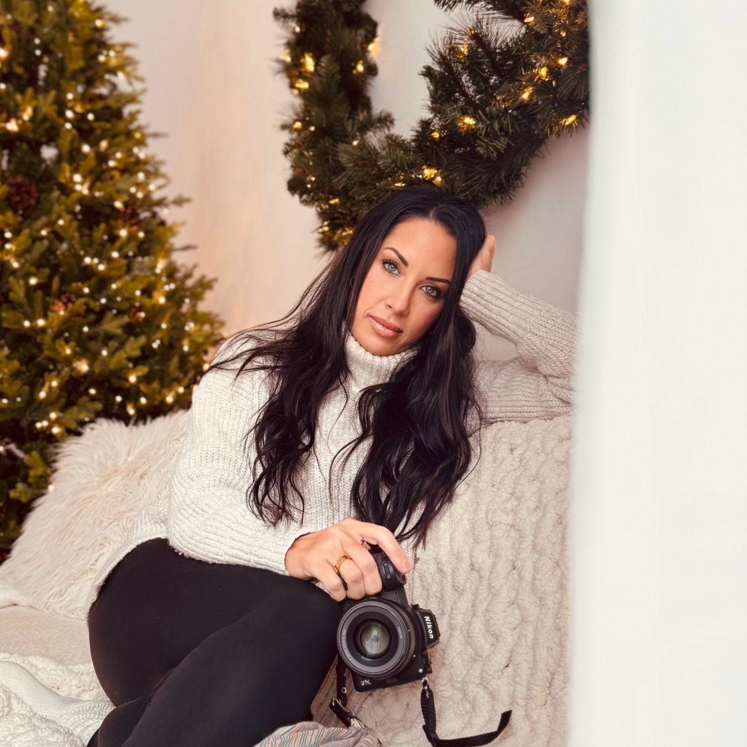 Dark-haired woman holding a camera sitting near decorated Christmas trees in a cozy setting.