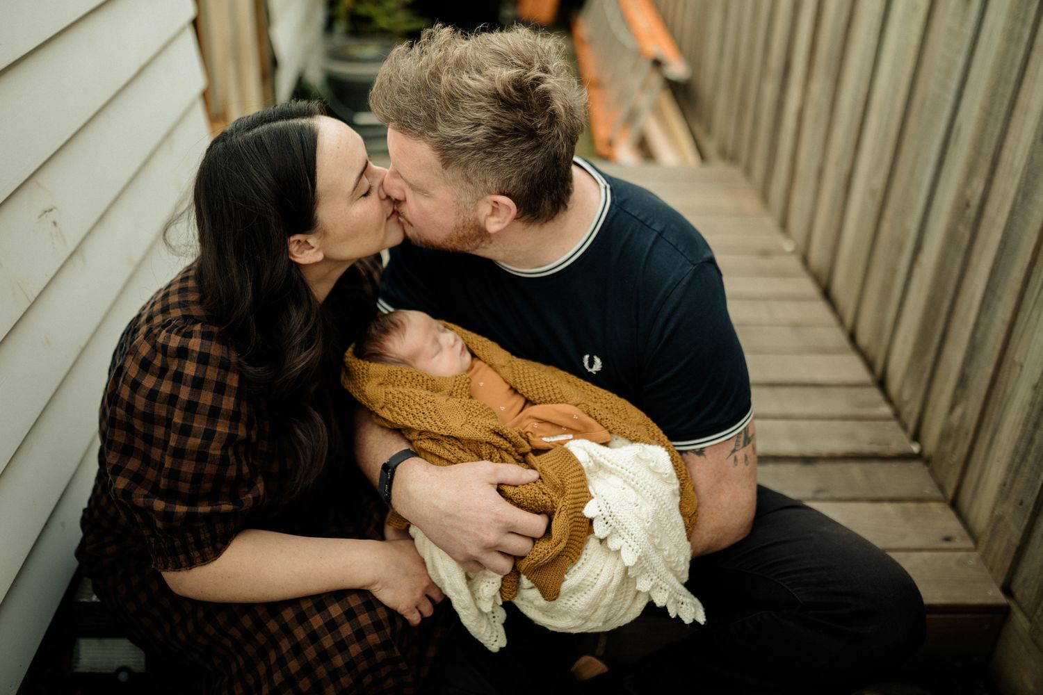 A couple shares an intimate moment while holding their baby wrapped in a mustard yellow blanket on a wooden walkway.