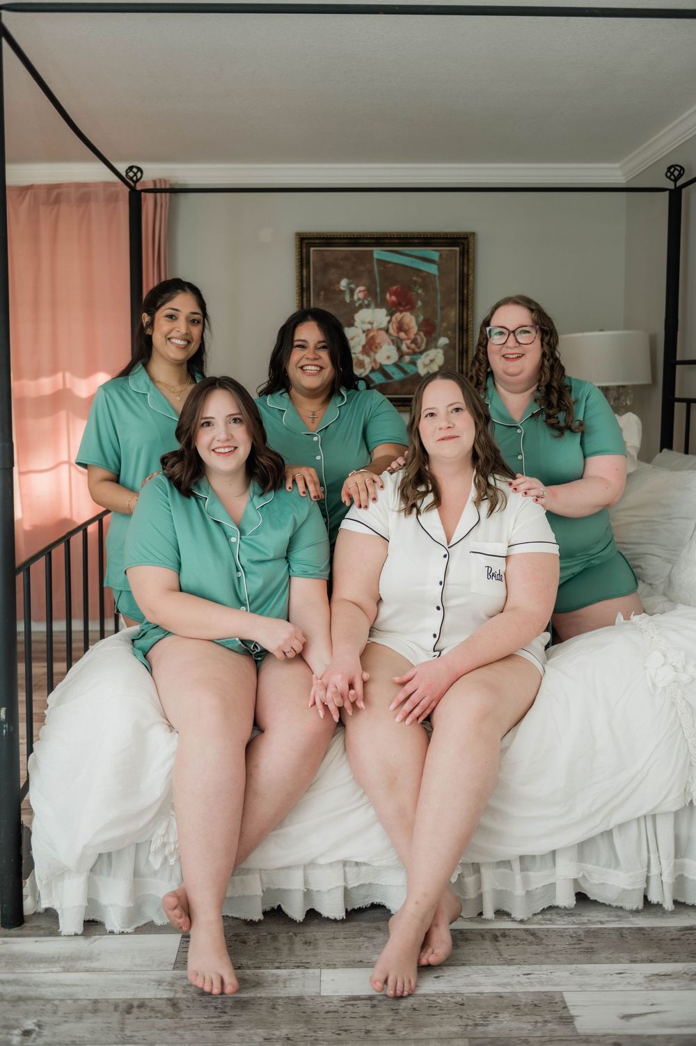 Group wearing matching teal robes and white pajamas sit together on white bedding for candid pre-wedding photo.