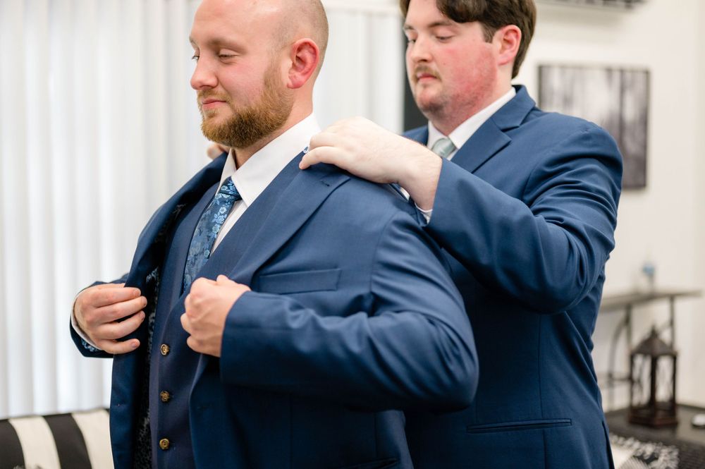 A groomsman helps adjust another's navy blue suit jacket during wedding preparations.