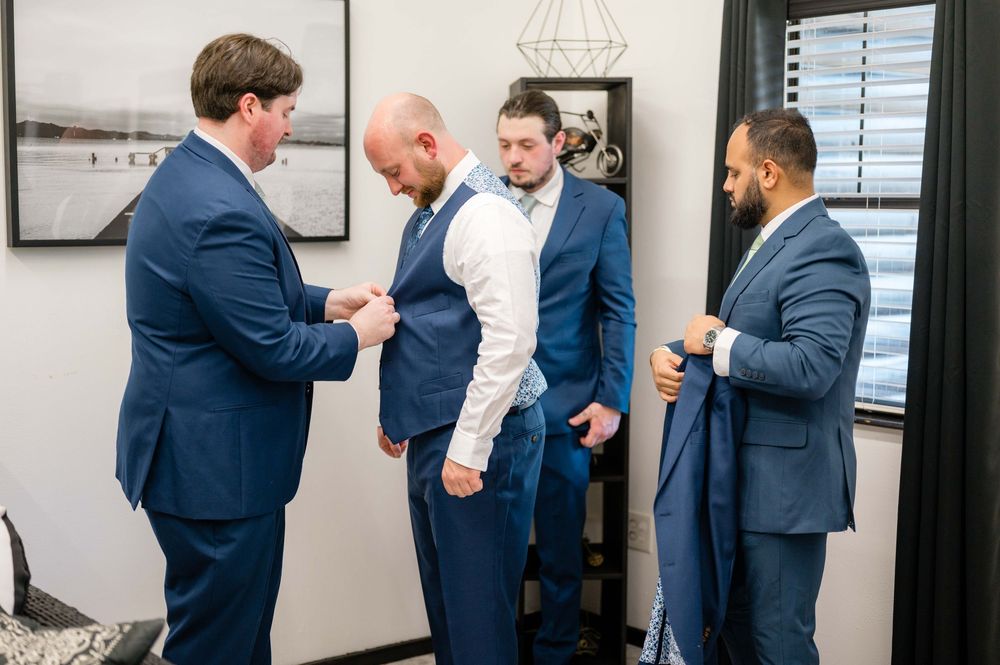 Groomsmen in navy suits help each other get dressed before the ceremony.