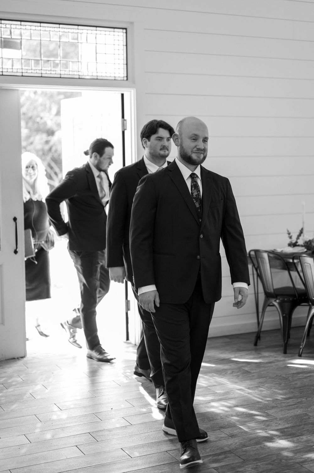 Three groomsmen in black suits walk together in a bright indoor venue during a wedding.