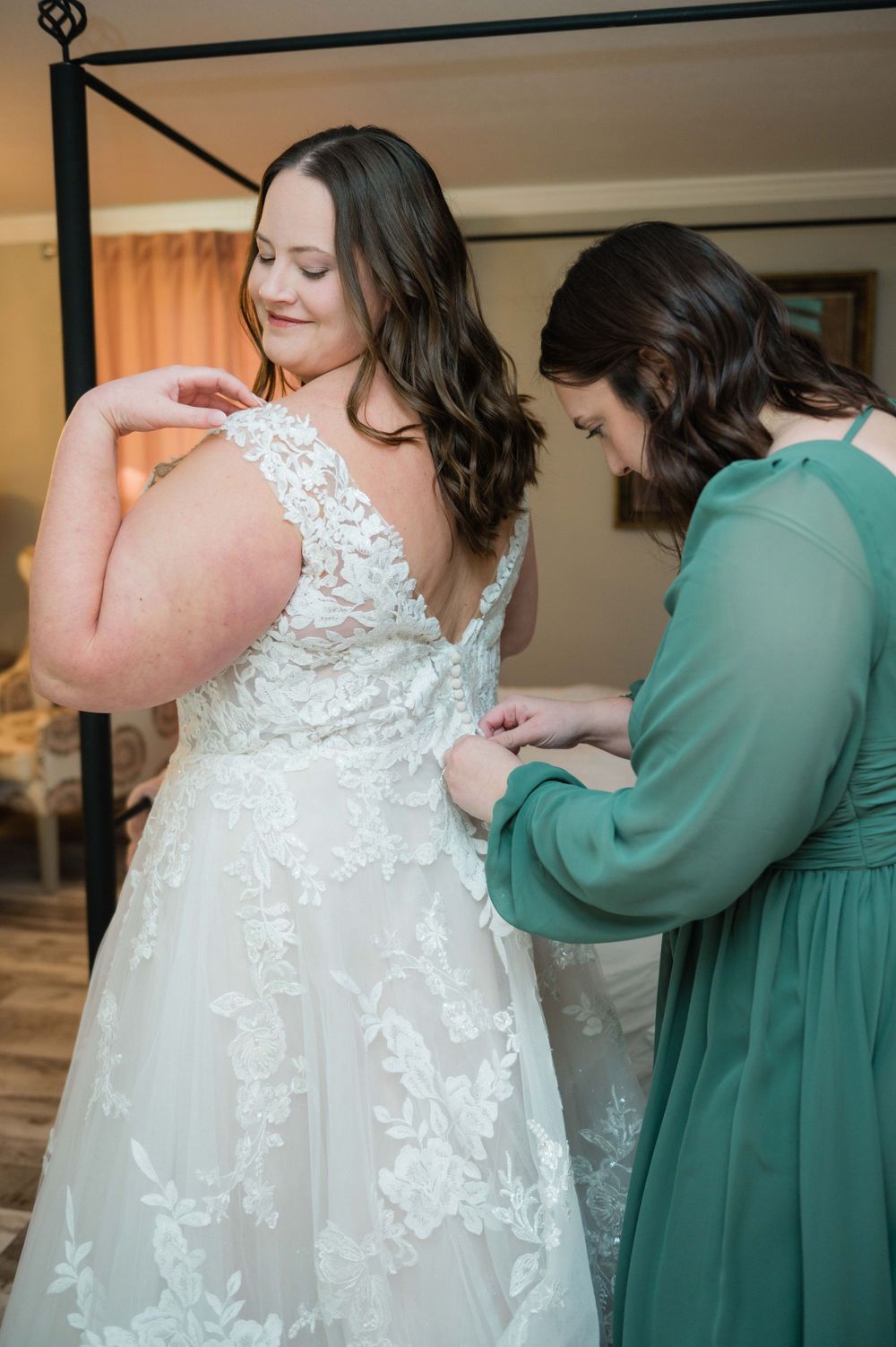 A bride in a lace wedding gown gets help with her dress from bridesmaids wearing sage green dresses.