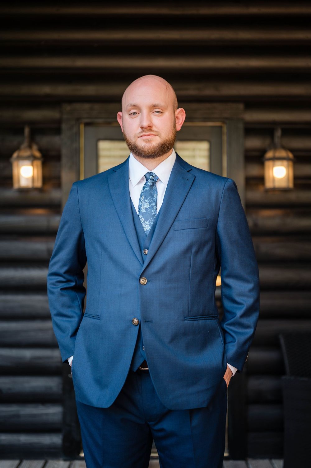 Professional portrait of someone in a navy blue suit and floral tie standing in front of a rustic log cabin with lanterns.
