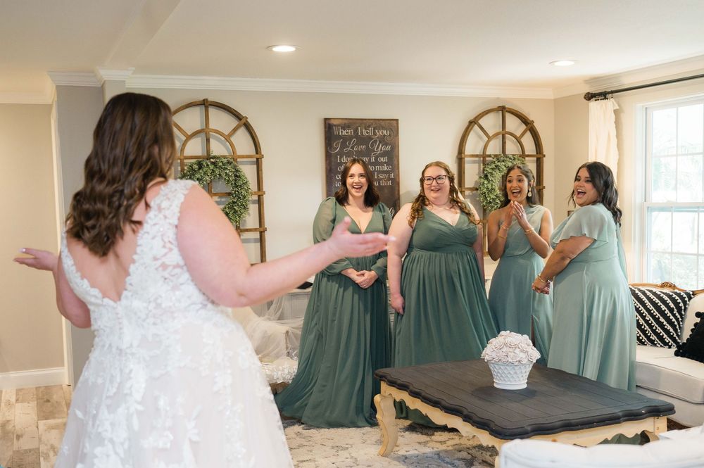 A bride in a white lace dress shares a joyful moment with bridesmaids wearing sage green dresses in an elegant indoor setting.