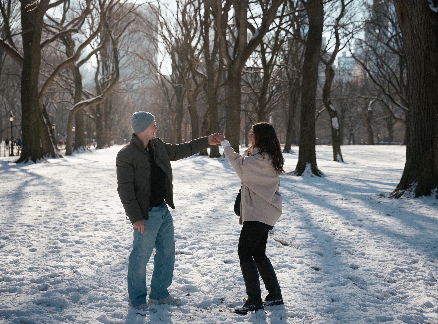A playful couple dances together in a snow-covered park surrounded by bare winter trees.