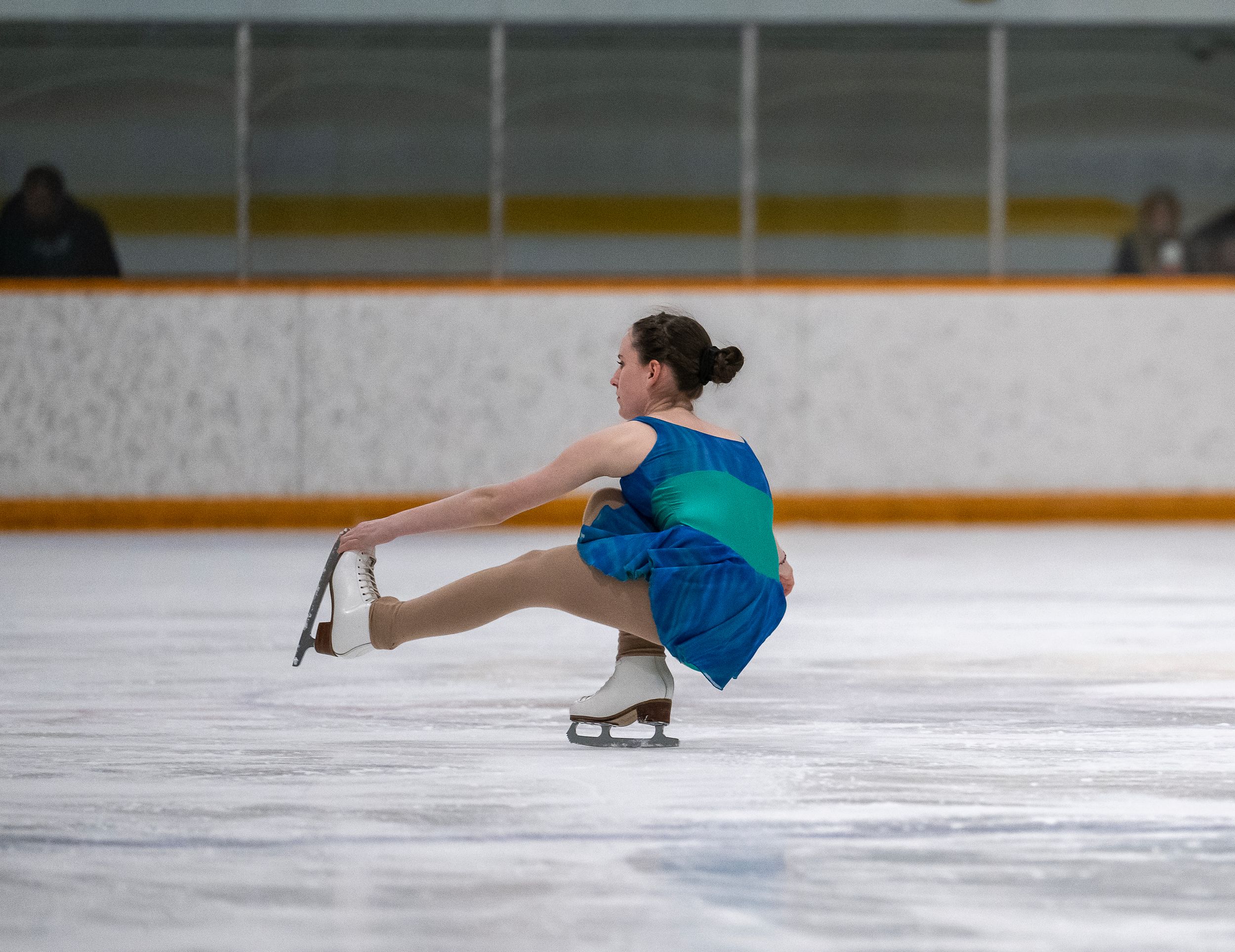 Figure skater performs a crouching spin on ice wearing blue and turquoise costume during practice session.