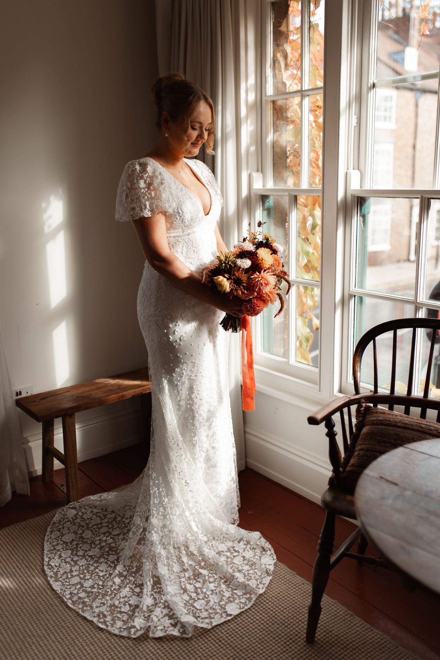 Elegant bride in lace wedding gown holds autumn-colored bouquet while standing by sunlit window in rustic room.