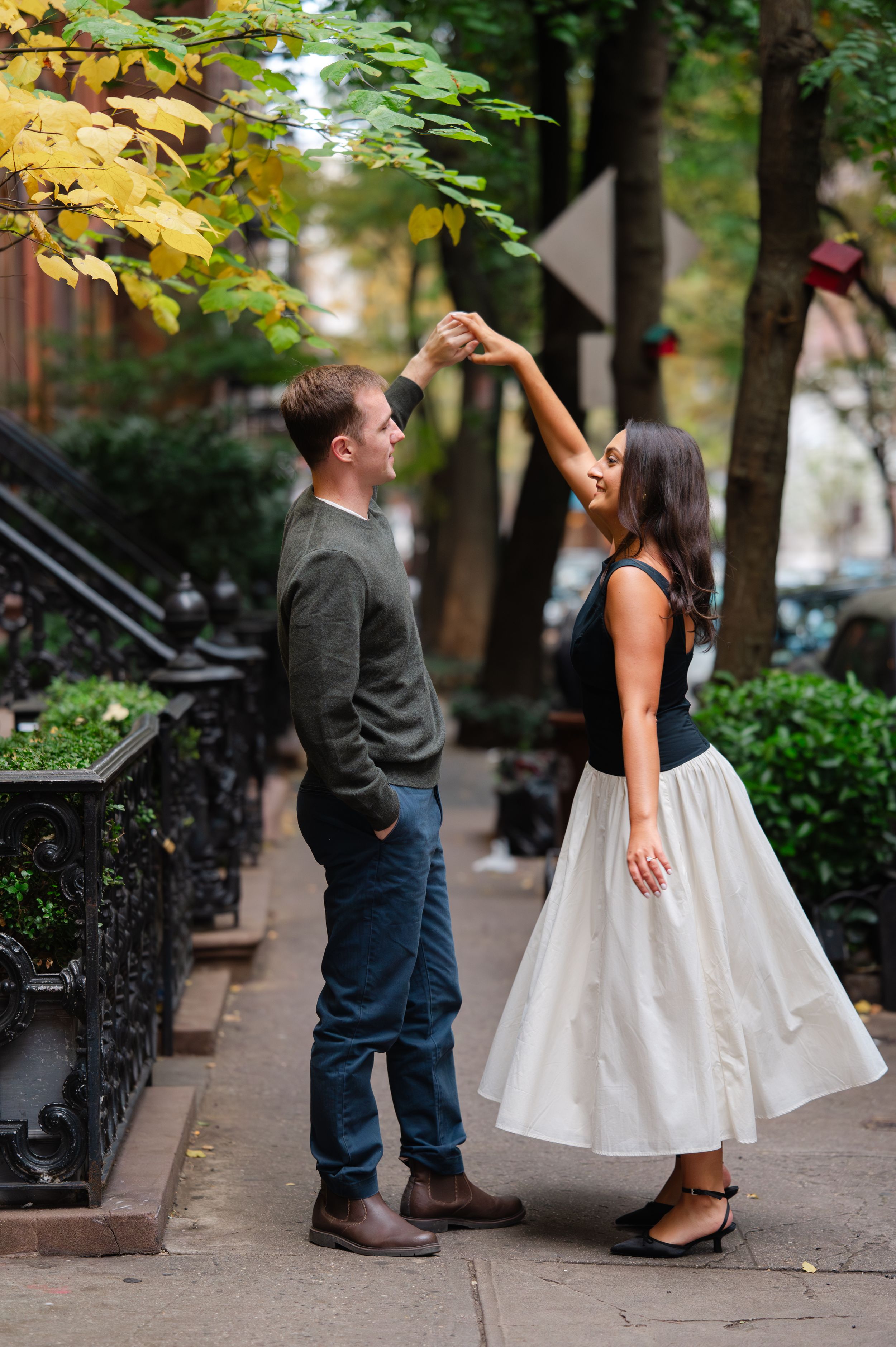 A couple in casual attire shares a romantic moment on a tree-lined urban sidewalk during an autumn day.