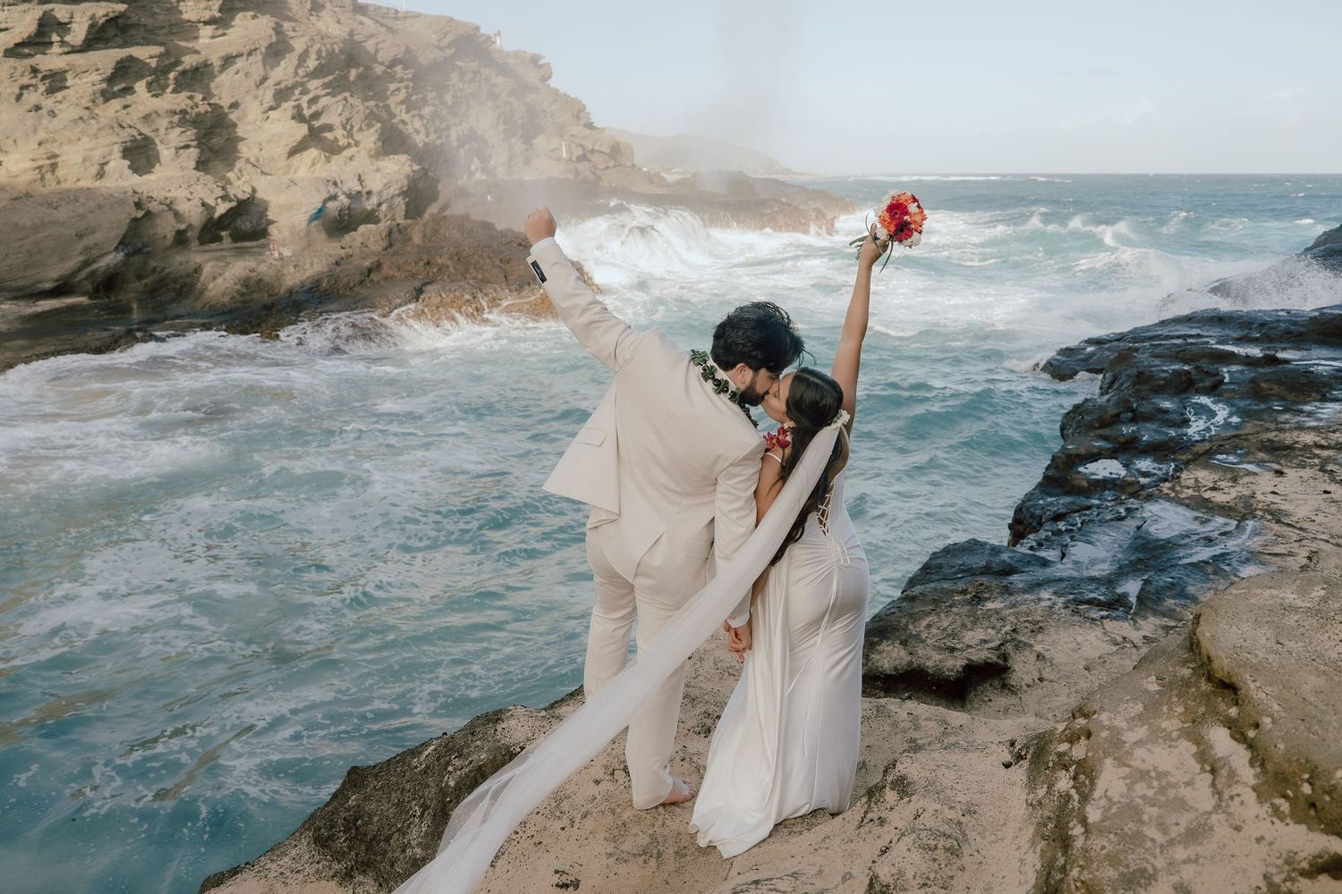 A couple in wedding attire stands on dramatic rocky cliffs overlooking crashing ocean waves at sunset.