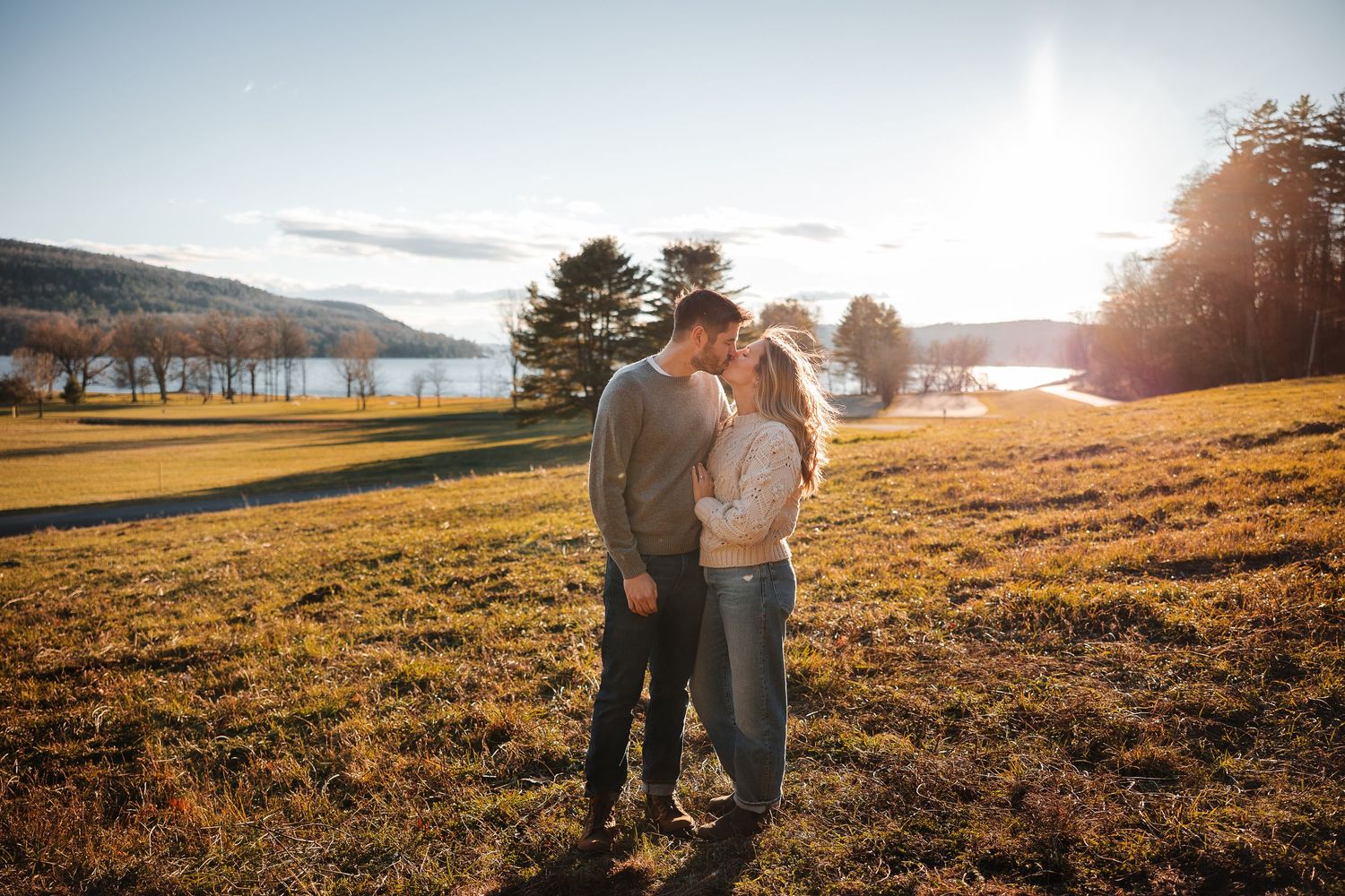 A couple embraces in a golden field at sunset with warm sunlight streaming through the trees.