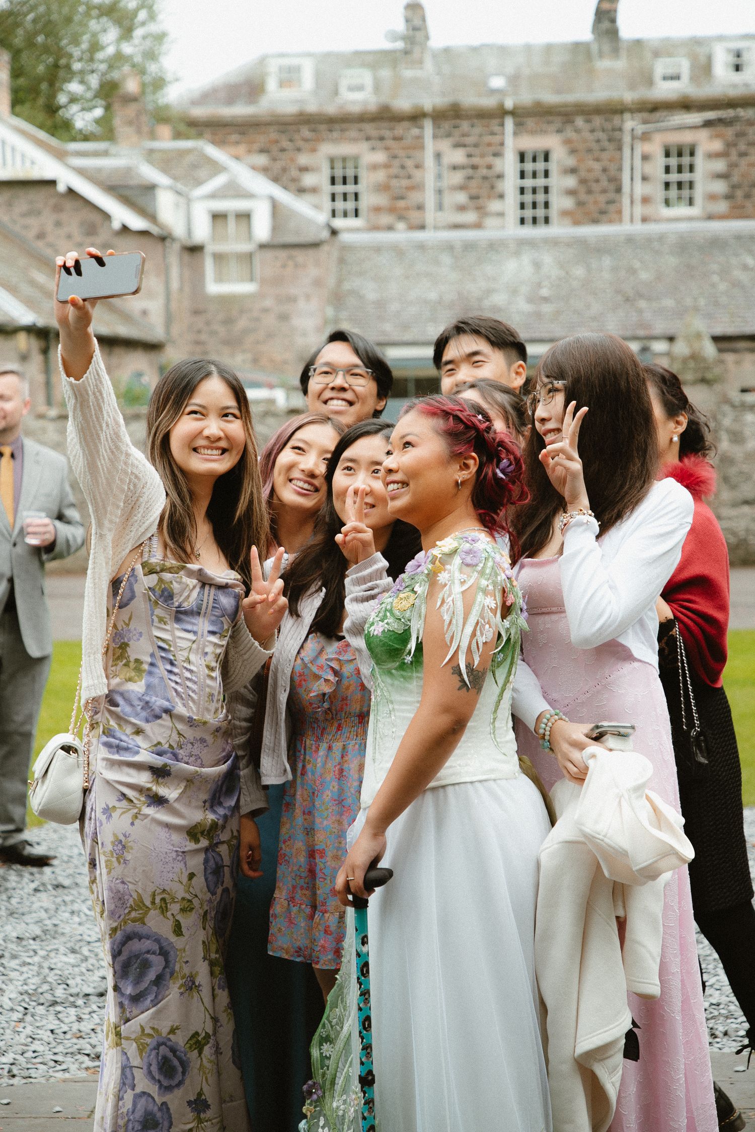 Group of wedding guests in colorful dresses laugh and celebrate outdoors near stone buildings.