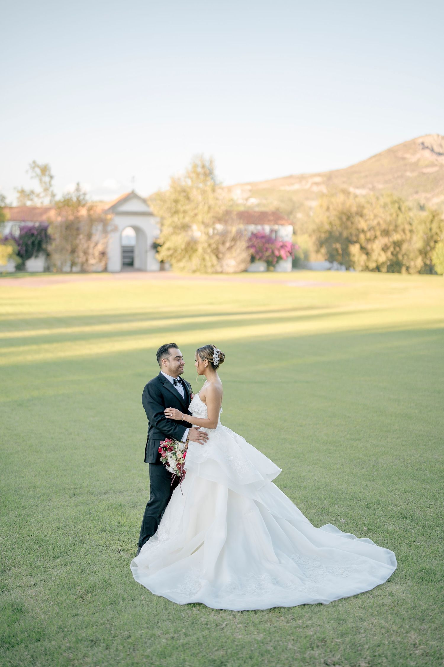 A couple in wedding attire dance together on a grassy field during golden hour with rolling hills in the background.