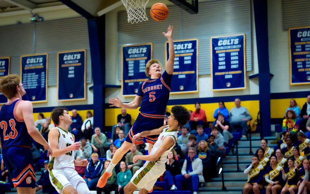 Hershey Trojans player jumps for a layup with the crowd and banners visible in the background by SportsFox Photography, Hershey PA.