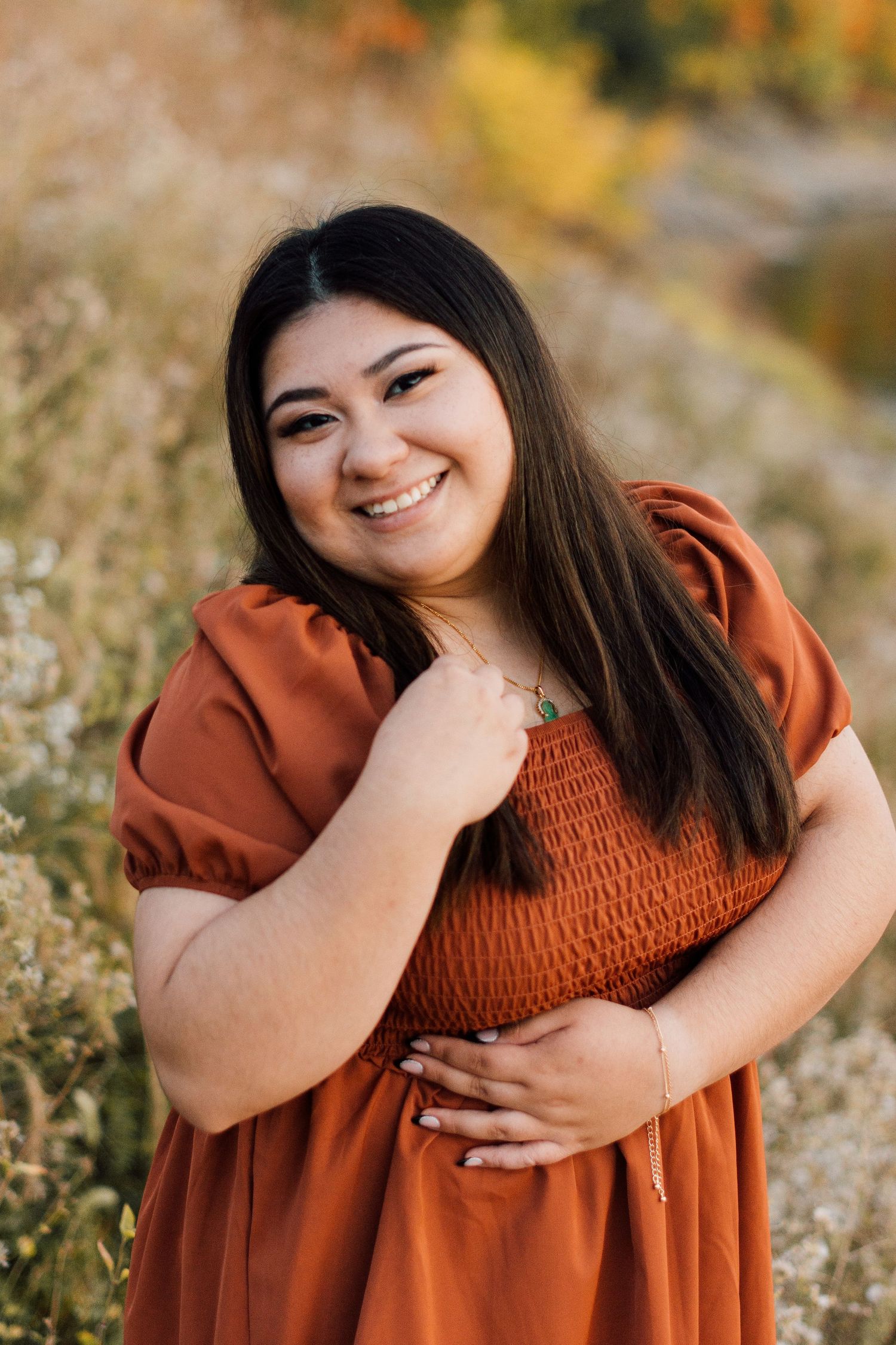 Woman wearing rust-colored dress smiles warmly in outdoor nature setting.