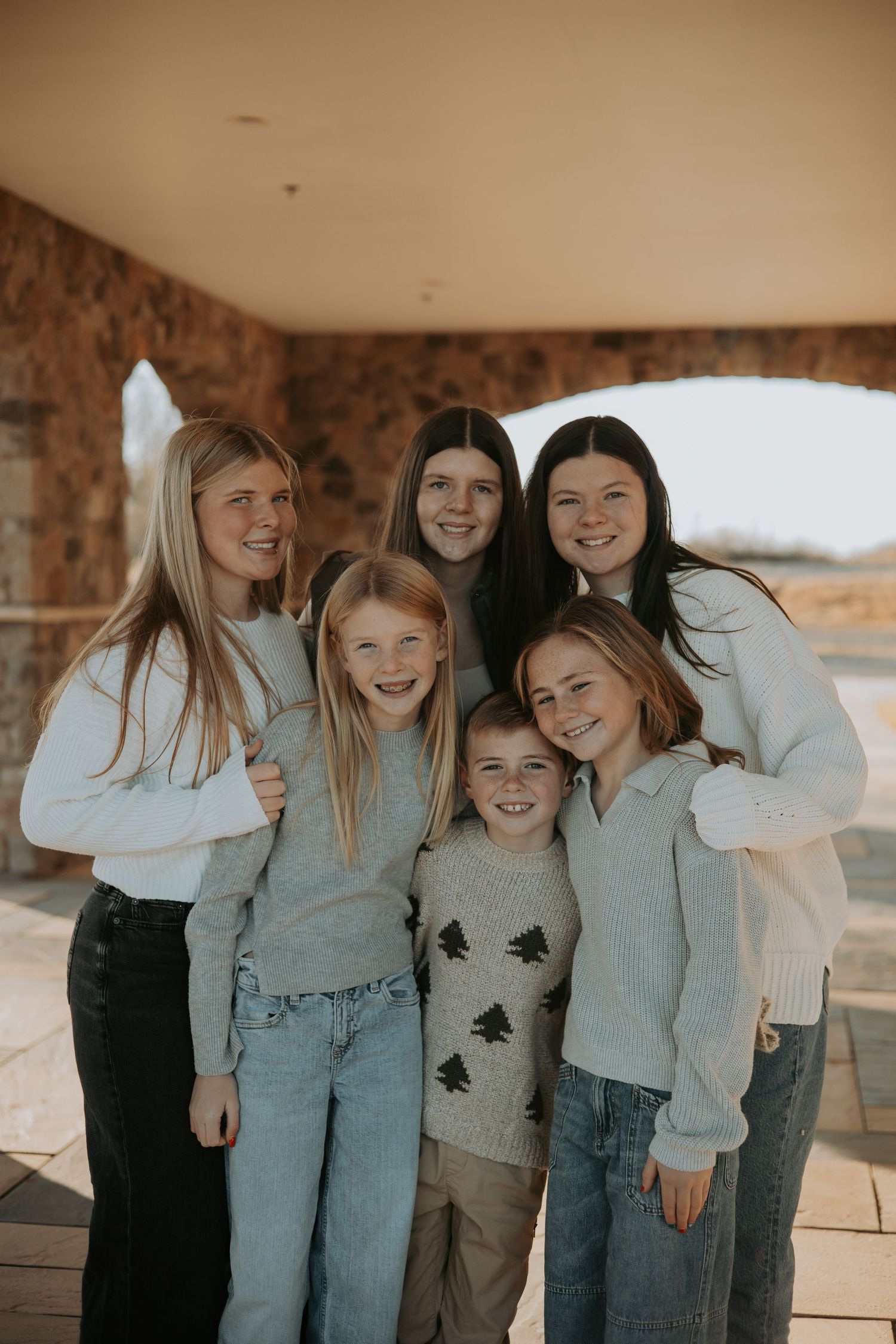 A group of six people in casual clothing stand together smiling on a covered porch in warm lighting.