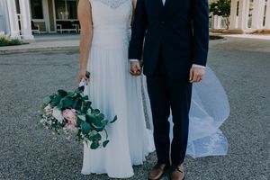Bride and groom hold hands outdoors, bride carrying a lush greenery and pink floral bouquet in a flowing white gown.