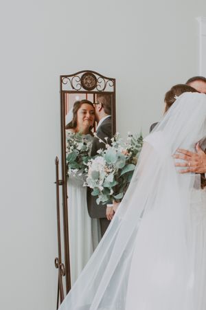 Bride and groom embrace in a mirror reflection, bridesmaid holding white and greenery bouquet nearby.