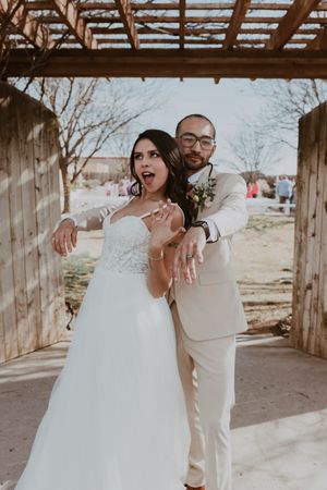 Excited bride and groom show off wedding rings under a rustic wooden pergola, groom in beige suit.