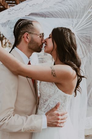 Bride and groom kissing under flowing white veil, groom in tan suit with braided hair and glasses.