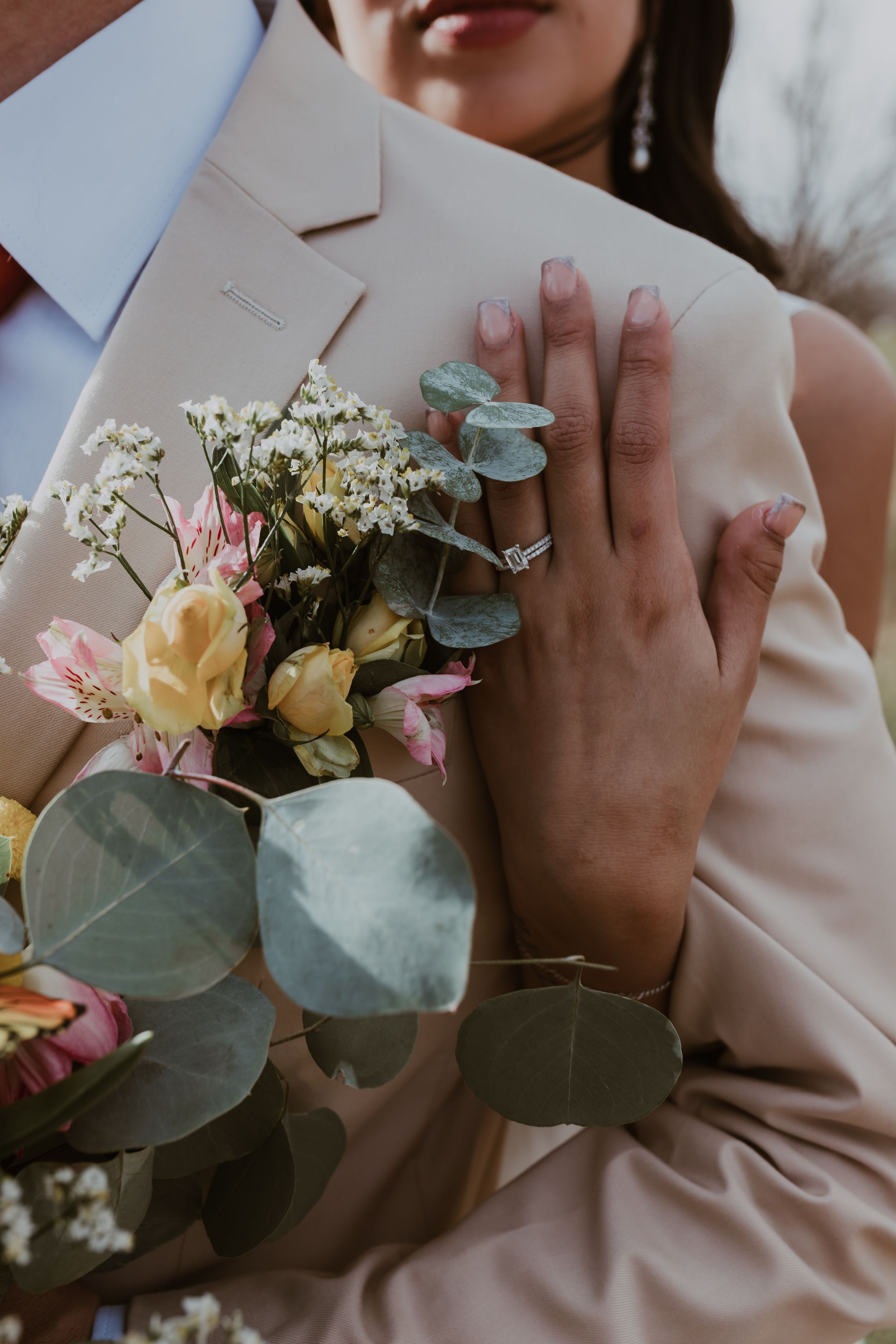 Close-up of bride resting hand on grooms tan suit lapel, diamond engagement ring visible beside floral boutonniere.