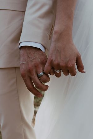 Close-up of newlyweds holding hands showing matching wedding bands, bride in white gown groom in tan suit.