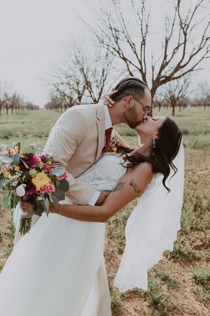 Bride and groom sharing passionate kiss outdoors, groom lifting bride while she holds vibrant floral bouquet.