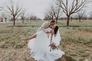Groom dipping bride in romantic kiss among bare trees, bride holding colorful wedding bouquet outdoors.