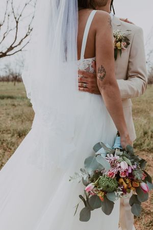 Close-up of bride and groom embracing from behind, bride holding vibrant bouquet with tattoo visible on arm.
