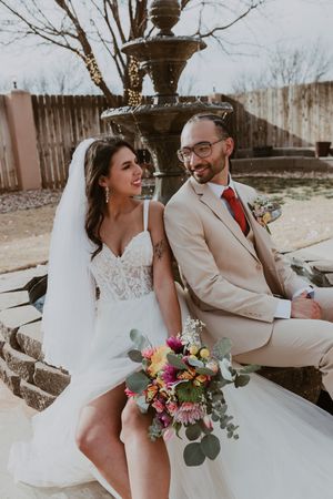 Smiling bride and groom sitting by stone fountain, bride holding colorful bouquet, groom in tan suit with red tie.