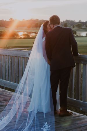 Bride and groom share a tender kiss on a wooden deck at sunset, bride's cathedral veil flowing in the breeze.