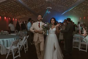 Bride and groom make a joyful grand entrance into a tented wedding reception with string lights overhead.