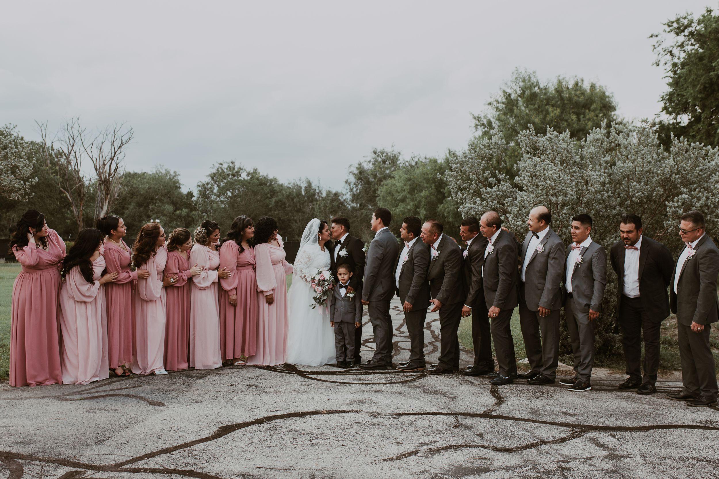 Wedding party poses outdoors with bridesmaids in pink dresses and groomsmen in gray suits flanking the bride and groom.