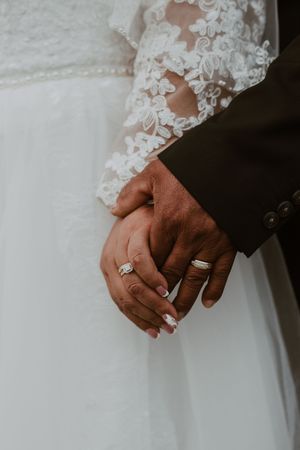 Close-up of bride and groom holding hands showing wedding rings, bride in lace gown and groom in dark suit.