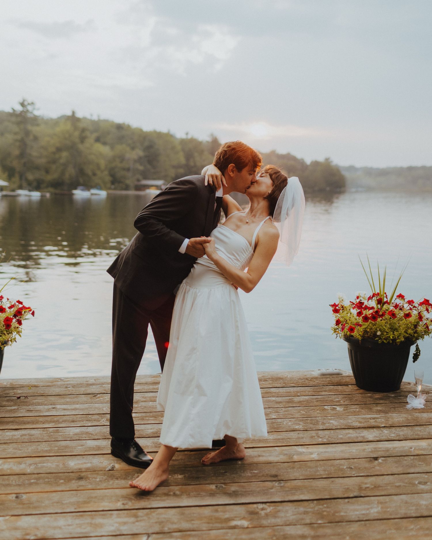A romantic couple shares a kiss on a wooden dock by a serene lake during a golden hour photoshoot.