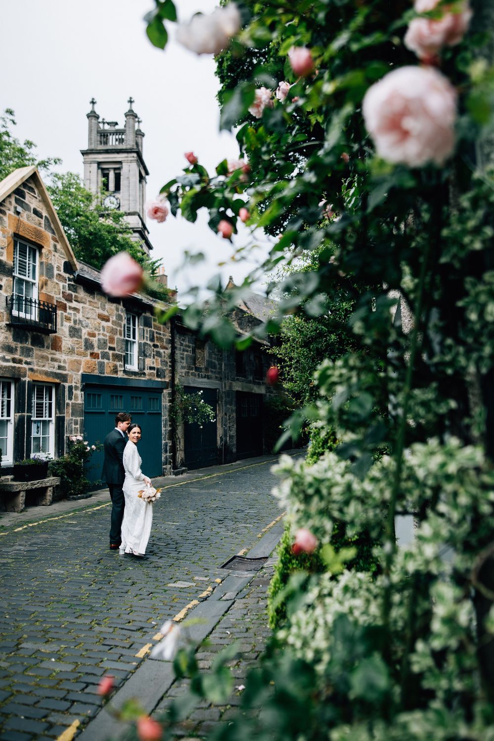A romantic scene on a cobblestone street in Edinburgh's historic district framed by blooming pink roses and stone buildings.