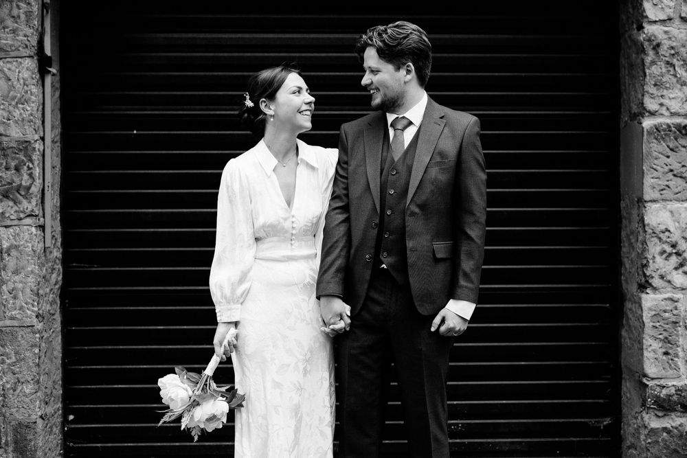 A black and white wedding photo of a couple standing together against a dark door, with the bride holding a bouquet.