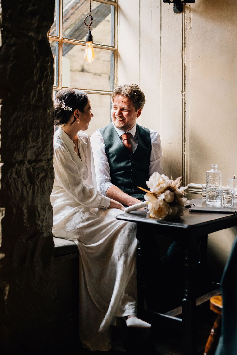 A couple shares an intimate moment at a small table by a window with vintage lighting and floral decor.